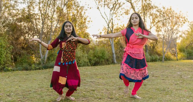 An image of two people performing a dance outdoors on grass, with trees in the background and arms extended in coordinated poses