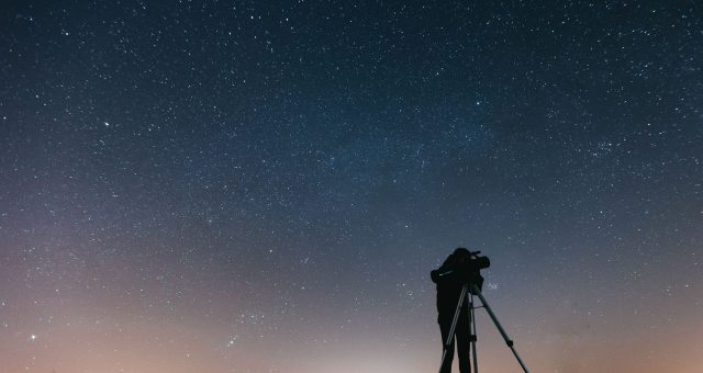 An image of a Silhouette of a person looking through a telescope on a tripod beneath a clear, star-filled night sky.