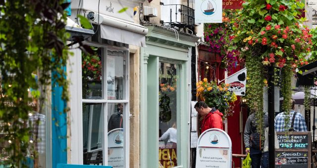 Pedestrian shopping street lined with independent shops, colourful hanging flower baskets and outdoor signs, with wet pavements reflecting the surrounding buildings.