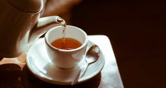 Tea is being pred from a white teapot into a white teacup and saucer with a spoon in the saucer