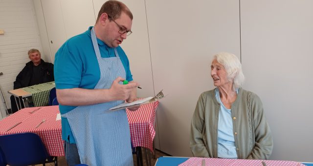 Person in a blue apron writes on a clipboard while standing beside a seated person at a table in a community hall.